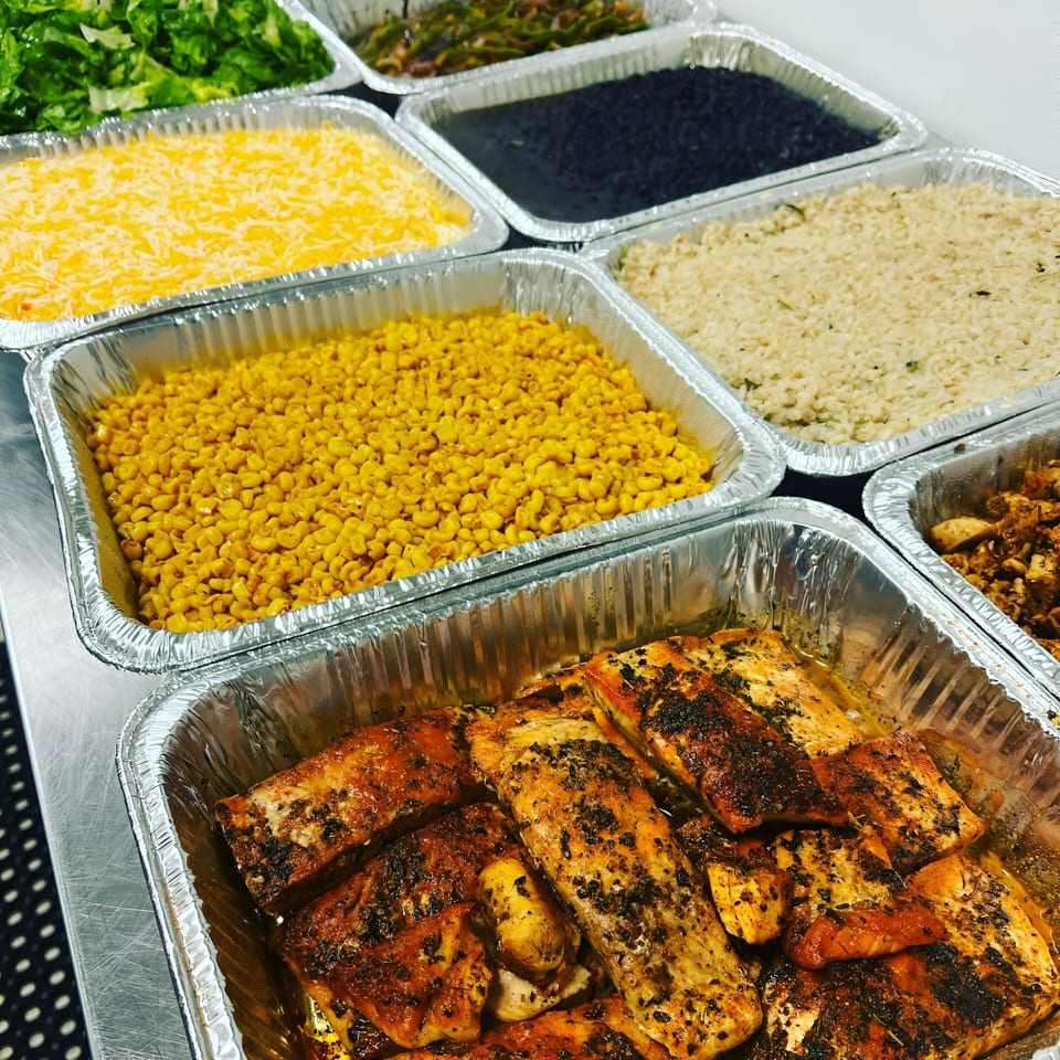 Healthy catering spread with salmon, corn, rice, black beans, cheese, and lettuce in foil trays.